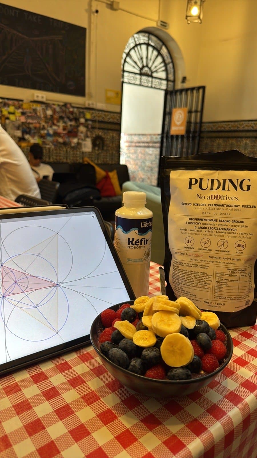 Stół w hostelu: tablet, kefir, paczka PUDING i miska z owocami