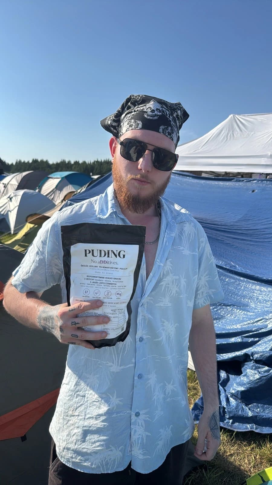 Klient w chuście i okularach przeciwsłonecznych trzyma paczkę Puding na polu namiotowym