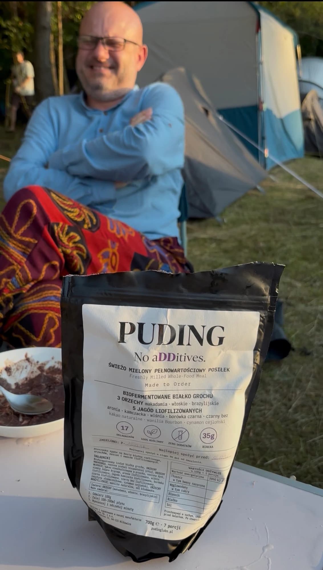 Paczka Puding na stole przed namiotem, za nią uśmiechnięty klient i pusta miska
